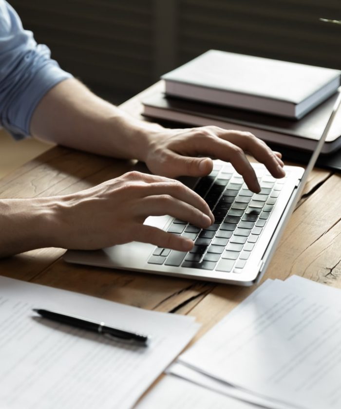 Close up businessman using laptop, typing on keyboard, sitting at wooden desk with documents, writing email, accountant writing financial report, busy student studying online, searching information